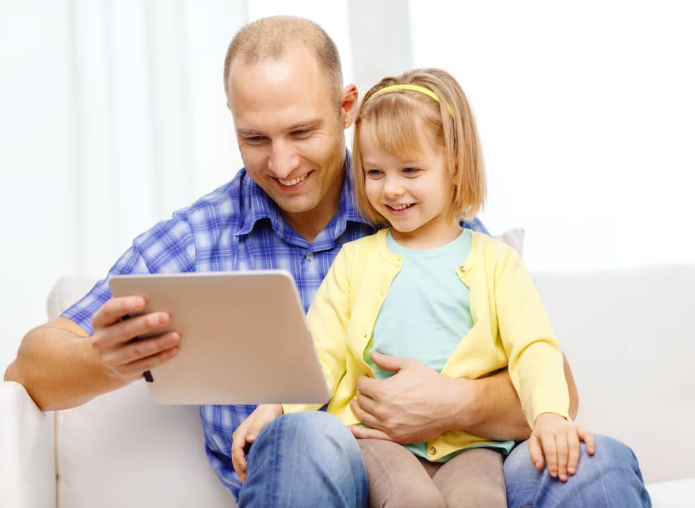 a father and daughter smile together as they watch a video on their tablet